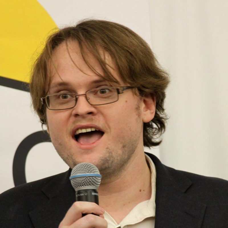 White man with medium length hair and rectangular glasses speaks into a microphone