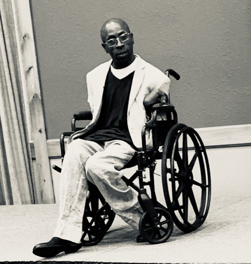 Black man in rectangular glasses and a white sports coat sitting in a wheelchair